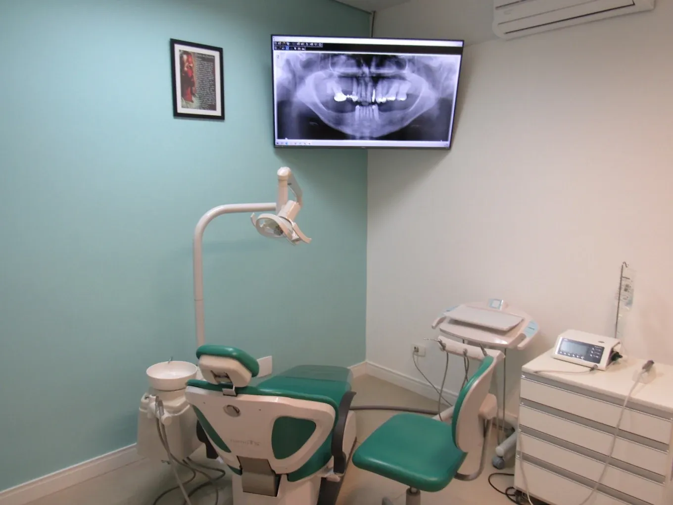 Consultório com cadeira odontológica verde e uma tela na parede para visualizar exames