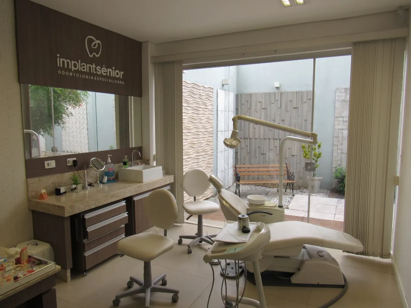Consultório com cadeira odontológica e uma janela de vidro com vista para o jardim